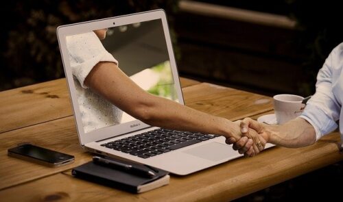 Auf einem Tisch steht ein Laptop. Aus dem Bildschirm des Laptops heraus ragt ein Arm und schüttelt der Person vor dem Laptop die Hand. Das Bild symbolisiert hier die Zusammenarbeit von der Person am einen oder der Person am anderen Ende der Verbindung.